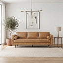 Mid-century modern tan genuine leather three-seater sofa with a tufted bench cushion, bolster pillows, and tapered wood legs in a bright living room with a potted olive tree, framed abstract art, brass chandelier, and side table.
