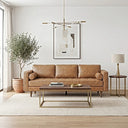 A mid-century modern tan leather sofa with bolster pillows and a tufted bench seat in a bright living room, flanked by a potted olive tree and a side table, behind a wood and brass coffee table.
