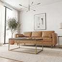 Mid-century modern tan genuine leather three-seater sofa with bolster pillows and wooden legs in a bright living room, styled with a wood-and-brass coffee table, cream woven rug, potted olive tree, framed abstract art, and a Sputnik chandelier.

