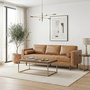 Mid-century modern tan leather sofa with bolster pillows in a bright living room, paired with a wood and brass coffee table, cream area rug, potted tree by tall windows, framed geometric wall art, and a modern brass chandelier.
