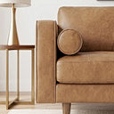 Close-up of a tan genuine leather sofa featuring a square track arm, cylindrical bolster pillow, thick seat cushion with stitched seams, and tapered wooden leg, set beside a brass-framed end table with a gold table lamp.

