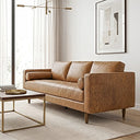 A mid-century modern tan genuine leather sofa with a tufted seat, three back cushions, two bolster pillows, and tapered wooden legs, set in a bright living room with a wood and brass coffee table, neutral area rug, and framed abstract art.
