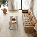 High-angle view of a tan genuine leather sofa with a tufted bench seat and bolster pillows in a bright living room, paired with a cream area rug, a wood-and-brass coffee table, and a potted tree by sheer curtains.
