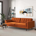 Casey tufted back burnt-orange velvet upholstered sofa with cylindrical bolster pillows and tapered wood legs, styled in a bright modern living room with wall art and an arched floor lamp.