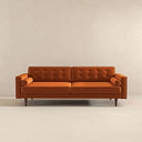 Casey mid-century modern burnt-orange velvet upholstered sofa featuring a button-tufted back, matching cylindrical bolster pillows, and tapered dark wood legs on a beige background.