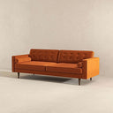 Three-quarters view of the Casey mid-century modern burnt-orange velvet upholstered sofa featuring a button-tufted back, matching cylindrical bolster pillows, and tapered dark wood legs on a beige background.