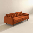 Three-quarters view of the Casey mid-century modern burnt-orange velvet upholstered sofa featuring a button-tufted back, matching cylindrical bolster pillows, and tapered dark wood legs on a beige background.