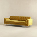 Three-quarters view of the Casey mid-century modern gold velvet upholstered sofa featuring a button-tufted back, matching cylindrical bolster pillows, and tapered dark wood legs on a beige background.