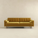 Casey mid-century modern gold velvet upholstered sofa featuring a button-tufted back, matching cylindrical bolster pillows, and tapered dark wood legs on a beige background.