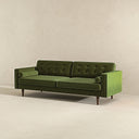 Three-quarters view of the Casey mid-century modern olive-green velvet upholstered sofa featuring a button-tufted back, matching cylindrical bolster pillows, and tapered dark wood legs on a beige background.