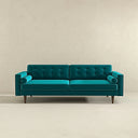 Casey mid-century modern teal velvet upholstered sofa featuring a button-tufted back, matching cylindrical bolster pillows, and tapered dark wood legs on a beige background.