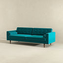 Three-quarters of the Casey mid-century modern teal velvet upholstered sofa featuring a button-tufted back, matching cylindrical bolster pillows, and tapered dark wood legs on a beige background.