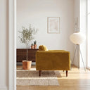 Side profile of the Casey gold velvet armchair with tapered wood legs in a bright, modern living room next to a potted olive tree, woven rug, and paper lantern floor lamp.