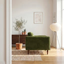 Side profile of a Casey olive green velvet armchair with tapered wooden legs styled in a sunlit modern living room with a potted olive tree and tripod floor lamp.