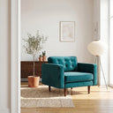 Casey teal velvet armchair with button tufting and tapered wooden legs styled in a modern, sunlit living room next to a potted olive tree.