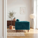 Side view of a Casey teal velvet armchair with tapered wood legs in a sunlit modern living room beside a potted olive tree and tripod floor lamp.