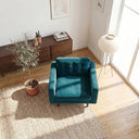 Casey teal velvet armchair with button-tufted back cushion on a woven rug in a sunlit living room with a potted olive tree and paper lantern floor lamp.