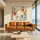 Chase 90-inch mid-century modern tan genuine top-grain leather sofa with tapered wooden legs, set in a bright, minimalist living room with an abstract painting on the wall and a floor-to-ceiling window overlooking the ocean.