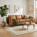 Chase 90-inch mid-century modern tan genuine top-grain leather sofa in a bright living room with a green throw blanket, round wooden coffee table, cream textured rug, and a potted fiddle-leaf fig tree.