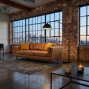 Chase 90 Mid-Century Modern tan genuine top grain leather sofa with yellow accent pillows in an industrial loft living room featuring exposed brick walls, large grid windows, a black arched floor lamp, and a city skyline view at dusk.