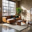 Chase 91-inch tan genuine leather left-facing sectional sofa in an industrial loft living room with exposed brick walls, large grid windows, dark grey knit throw, wooden coffee table, and cream rug.
