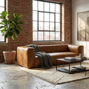 Chloe 92-inch tan genuine leather sofa with a grey knit throw blanket in a modern loft living room featuring exposed brick walls, a large industrial window with city views, a black coffee table, a potted fiddle-leaf fig, and a textured area rug.