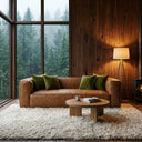 Chloe 92-inch tan genuine leather sofa with olive green velvet pillows in a modern living room featuring a wood-paneled wall, a round wooden coffee table on a shaggy area rug, a tripod floor lamp, and a large window looking out at a misty pine forest.
