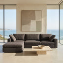 Concetta 121-inch dark gray fabric modular sectional sofa with chaise lounge in a sunlit modern oceanfront living room with floor-to-ceiling windows and a low travertine coffee table.