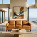 Cooper mid-century modern tan leather sofa in a sunlit coastal living room overlooking the ocean, with a travertine coffee table and abstract wall art.