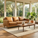 Cooper mid-century modern tan leather sofa in a sunlit living room with large windows, styled with a wooden coffee table on a textured cream area rug and a large potted Monstera houseplant.
