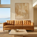 A tan leather three-seater reclining sofa with button-tufted back cushions and tapered wooden legs in a bright living room. A low square stone coffee table with a ceramic bowl rests on a woven rug in front of the couch. Sunlight streams in through large oceanfront windows, highlighting a neutral abstract painting hanging on the wall above the sofa.