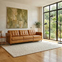 A three-seater tan leather electric reclining sofa with button-tufted back cushions in a sunlit modern living room. The sofa rests on a textured cream rug over light wood floors in front of a large abstract green painting. To the right, floor-to-ceiling black-frame windows look out onto lush garden greenery, with a potted tree and a black multi-arm floor lamp nearby.