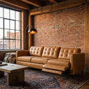 Three-seater Cora tan leather sofa with button-tufted back cushions and an extended right electric reclining footrest, set in an industrial loft with an exposed red brick wall, large gridded factory window, rustic wood coffee table, arched black floor lamp, and vintage patterned area rug.