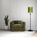 Olive green velvet swivel barrel chair with vertical channel tufting, a plush seat cushion, and a round gold base, set in a modern room with white paneled walls and light grey carpet. To the left is a potted snake plant in a white ceramic planter, and to the right is a slim black floor lamp with a matching green shade.