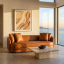 Dina 85-inch modern cognac leather sofa with matching throw pillows, set in a minimalist living room featuring a travertine stone coffee table, abstract warm-toned wall art, and large windows with a scenic ocean view at golden hour.