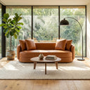 Dina 85-inch modern cognac leather sofa in a sunlit living room, styled with a round wooden coffee table, a potted fiddle-leaf fig, and an arched black floor lamp in front of large floor-to-ceiling windows.