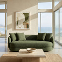 Dina 85 modern olive green boucle sofa with curved ends and matching throw pillows, set in a bright coastal living room with floor-to-ceiling windows overlooking the ocean. A travertine stone coffee table with a decorative bowl sits on a woven jute rug in the foreground.