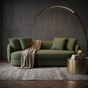 A Dina 85-inch modern olive green boucle sofa styled in a contemporary living room with a beige throw blanket, matching pillows, a minimalist black arc floor lamp, a brass drum side table, and a concrete wall background.