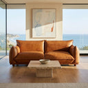 Elena 89 MCM cognac genuine leather sofa in a modern living room with floor-to-ceiling windows overlooking a coastal ocean view, styled with a square travertine stone coffee table and abstract art.