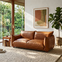 Elena 89-inch mid-century modern cognac genuine leather sofa in a sunlit living room. The couch is placed on a textured beige rug, flanked by a wooden side table with a brass lamp and a potted fiddle-leaf fig plant under a framed abstract art piece.