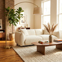 Elena 89-inch mid-century modern cream boucle sofa styled in a cozy living room with a walnut coffee table, dried pampas grass, arched brass floor lamp, textured beige area rug, and a potted fiddle leaf fig tree near an exposed brick wall.