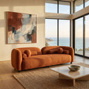 A modern Elissa 93-inch Japandi burnt orange boucle sofa with matching round and bolster pillows in a sunlit living room with floor-to-ceiling windows showing a coastal ocean view, an abstract painting on the wall, and a stone coffee table on a jute rug.