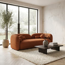 Elissa 93-inch Japandi burnt orange boucle sofa featuring a curved, low-profile design and matching spherical pillows in a minimalist living room. The modern rust-colored couch is set on a cream shag rug next to a dark wood coffee table and a potted tree, bathed in natural light from large black-framed glass doors.