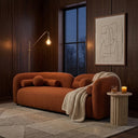 Modern Japandi-style Elissa 93-inch burnt orange bouclé sofa in a cozy living room with dark wood paneled walls. The sofa is styled with matching round and bolster pillows, a cream knit throw blanket, and sits on a geometric patterned rug. Beside it, a fluted travertine side table holds a lit candle and a drink next to an arched brass floor lamp, with abstract line art on the wall and a window showing twilight.