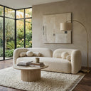 Elissa 93 Japandi ivory boucle sofa with round and bolster pillows in a minimalist living room, featuring a ribbed wood coffee table, a textured cream shag rug, a brass arch floor lamp, abstract wall art, and a large black-framed corner window with a garden view.