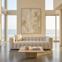 Elizia 86-inch Modern Chesterfield cream boucle sofa with channel tufting in a minimalist, sunlit living room. The low-profile cream couch is decorated with a matching boucle throw pillow and placed in front of a square travertine coffee table, with a large neutral abstract painting on the wall behind it and floor-to-ceiling windows showing a beach and ocean view.