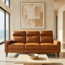 Elton 79-inch incliner sofa in waxy tan leather, shown in a modern, sunlit living room with large windows overlooking the ocean, an abstract painting on the beige wall, and a stone coffee table in the foreground.