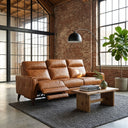 Elton 79 waxy tan leather left-facing incliner sofa with one seat extended, styled in an industrial loft living room with exposed brick walls, a rustic wooden coffee table, a black arched floor lamp, and a potted plant.