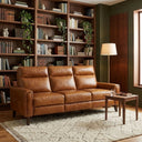 Elton 79 incliner sofa in waxy tan leather, shown in a cozy living room setting with a large wooden bookshelf, cream area rug, wooden side table, and floor lamp.