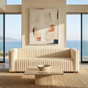 Enno 91-inch mid-century modern cream velvet sofa featuring vertical channel tufting, styled in a bright coastal living room with a square travertine coffee table, woven jute rug, abstract wall art, and floor-to-ceiling windows overlooking the ocean.